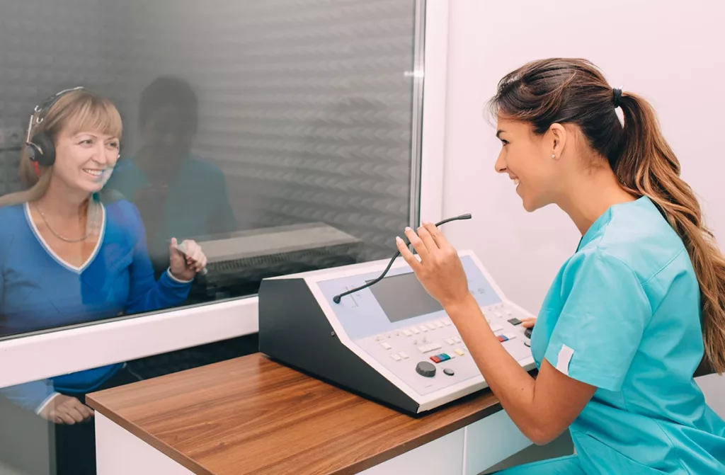 An older adult women in a sound booth at a hearing clinic happily undergoing her yearly hearing test as recommended by her hearing care specialist.